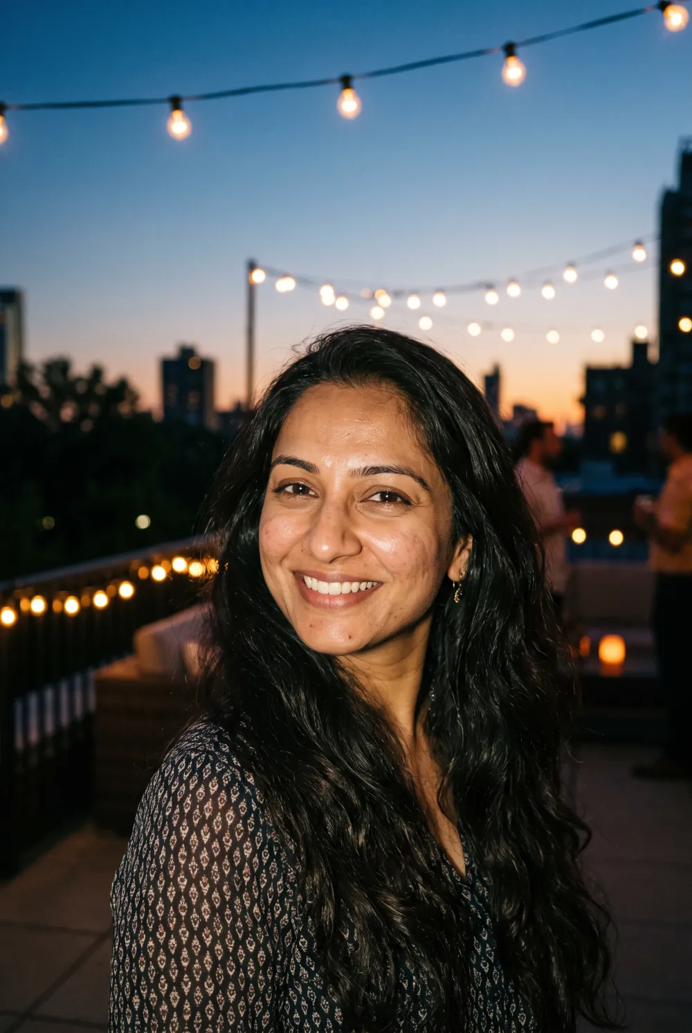 Smiling woman with long dark hair on a rooftop terrace at dusk under string lights, lit by a soft flash